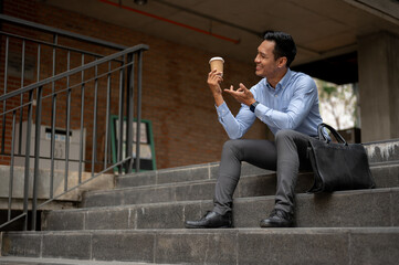 A handsome millennial Asian businessman enjoying his coffee on the stairs in front of the building.