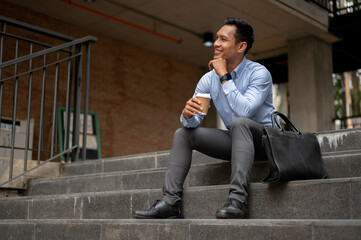A handsome millennial Asian businessman enjoying his coffee on the stairs in front of the building.