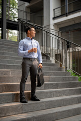 A handsome, confident Asian millennial businessman stands on the stairs outside of the building.
