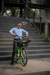 An exuberant Asian millennial businessman joyfully rides a green bicycle down stairs.