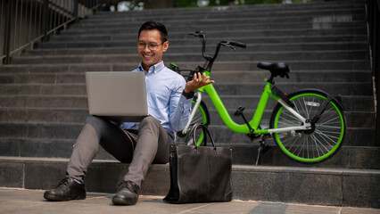 A confident Asian businessman sits on steps, working on a laptop with a smile.