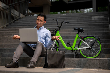 A confused Asian businessman sits on steps with a laptop, showing puzzled expression.