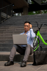 A determined Asian businessman sits on steps, working on a laptop, deep in thought.