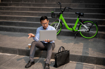 A confused Asian businessman sits on steps with a laptop, showing puzzled expression.