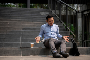 An Asian businessman sits on outdoor steps with a laptop and a briefcase, expressing confusion.