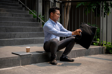 A stressed Asian businessman sits on outdoor steps, opening or finding something in his briefcase.