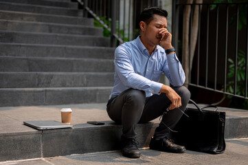 An Asian businessman sits on outdoor steps, experiencing frustration and the fear of being fired.