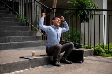 An angry Asian businessman sits on outdoor steps, holding a laptop with frustration, being fired.