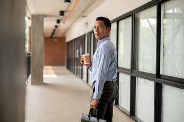 Determined Asian businessman walking along an indoor corridor, holding a coffee cup and a briefcase.