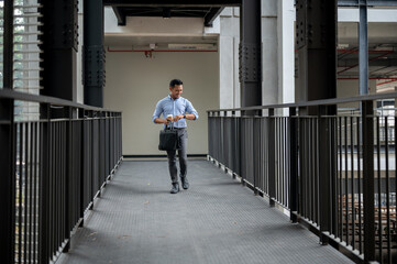 A confident Asian businessman is checking time on his wristwatch while walking along a corridor.