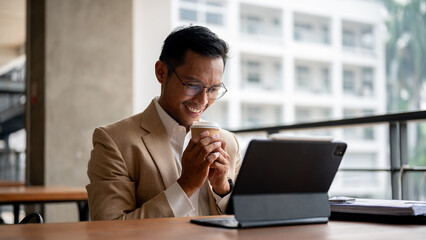 A happy Asian businessman smiles as he holds a disposable coffee cup while looking at a tablet.