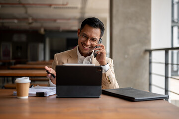 A confident businessman sits at a wooden table with a tablet and documents, talking on the phone.