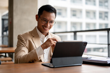 A confident Asian businessman sits at a table, working on his digital tablet and document.