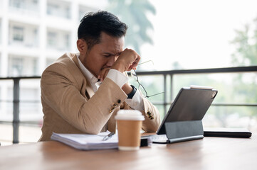 A frustrated Asian businessman covers his face with his hands while looking at a digital tablet.