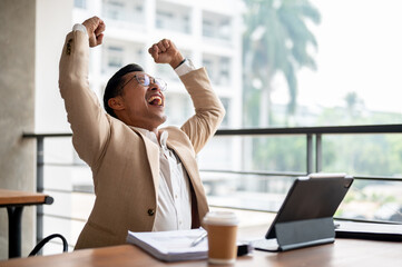 A cheerful Asian businessman raises his arms triumphantly, celebrating success or good news. - 781922860