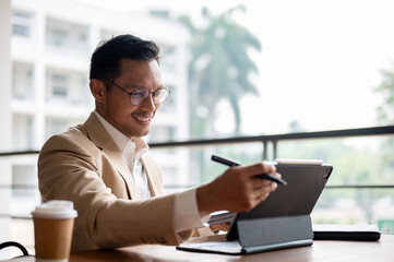 A confident Asian businessman sits at a table, working on his digital tablet and document.