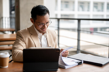 A confident Asian businessman is reviewing documents and working on a digital tablet.