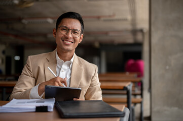 A confident Asian businessman sits at a table in a building corridor with his tablet and document.