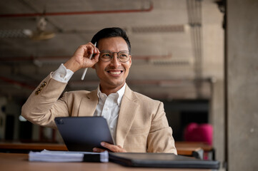 Happy Asian businessman smiles while holding a digital tablet and adjusts his glasses with one hand.