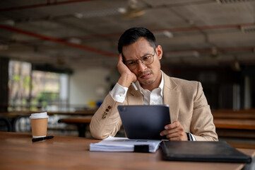 A contemplative Asian businessman looks pensive while holding a tablet, facing a problem at work.