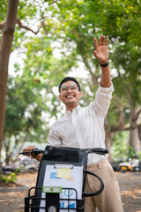 A happy businessman smiles and waves while pushing his bike, greeting someone while heading to work. - 781923295