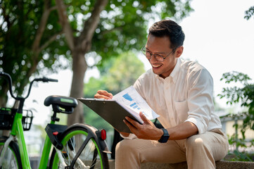 A confident, happy Asian millennial businessman reviewing his business document in a public park. - 781923410