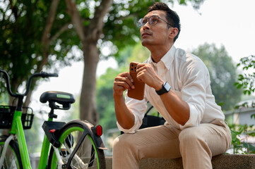 Pensive Asian businessman sits on a bench in a park, holding his phone, looking up, deep in thought.
