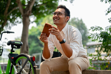 An optimistic Asian businessman sits on a bench outdoors, holding his phone with a smile.