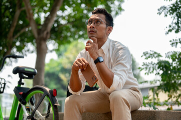 Pensive Asian businessman sits on a bench in a park, holding his phone, looking up, deep in thought.