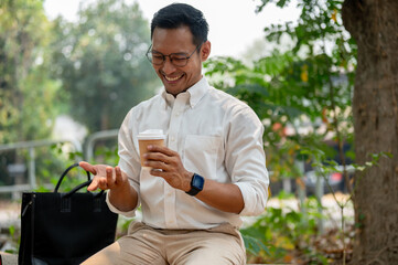 A happy Asian businessman sits outdoors, enjoying a coffee break in the public park on a bright day.