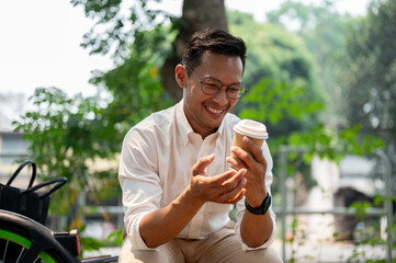 A happy Asian businessman sits outdoors, enjoying a coffee break in the public park on a bright day. - 781923638