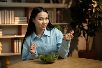 A woman is seated at a wooden table, savoring a bite of fresh green salad from a clear bowl in a...