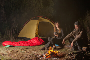 a man, a woman are sitting around a campfire in the evening in nature. There is a yellow tent and a...