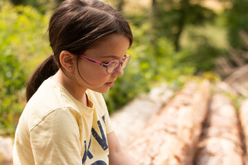 A small beautiful girl with glasses and a ponytail, smiling, sits on the sawn trunks of a pine...