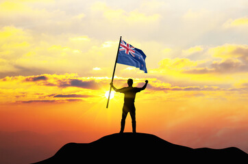 New Zealand flag being waved by a man celebrating success at the top of a mountain against sunset or sunrise. New Zealand flag for Independence Day.