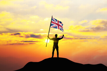 United Kingdom flag being waved by a man celebrating success at the top of a mountain against sunset or sunrise. United Kingdom flag for Independence Day.