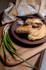 Clay plate of fried meat pies with cutlery and green onion on wooden table.