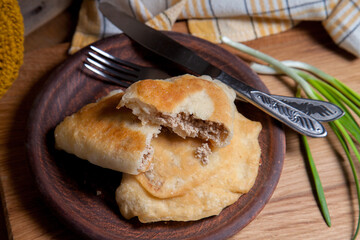 Clay plate of fried meat pies with cutlery and green onion on wooden table.
