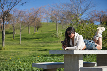 Fototapeta premium Young, beautiful, brunette woman in a white sweater, denim miniskirt and sunglasses, checking social media on her smartphone lying on a park bench on a sunny day. Beauty, fashion, trend concept.