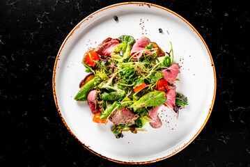 Top view of a gourmet salad with medium-rare sliced meat, fresh greens, and vegetables, artistically arranged on a white rustic plate against a black background