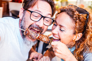 Happy adult couple have fun together eating meat cooked on a spit in touristic restaurant. Man and woman enjoy time eating meal from same fork posing for a picture. Happiness relationship food concept