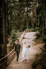 A bride and groom walk hand in hand down a forest path, with the groom leading and the bride...