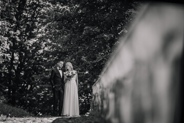 A black and white photo of a groom in a suit and a bride holding a bouquet standing together in a serene, leafy outdoor setting, evoking a sense of romance and elegance.