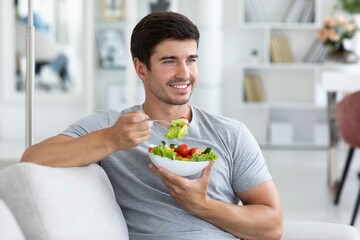 Happy man sitting on sofa at home while eating green vegetable salad. Healthy eating