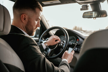 Surfing the internet by using smartphone. Man in black suit is sitting in the car, driving