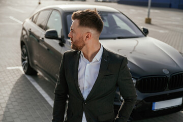 Focused portrait, against vehicle. Businessman in suit is near his black car outdoors