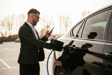 Holding wire for charging vehicle. Businessman in suit is near his black car outdoors