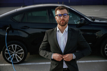 Handsome smart guy in glasses. Businessman in suit is near his black car outdoors
