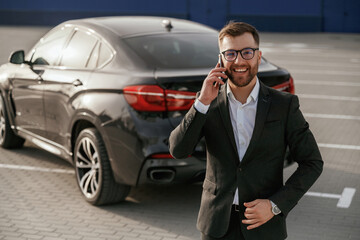 Cheerful facial expression. Businessman in suit is near his black car outdoors