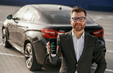 Charging process icon in the smartphone. Businessman in suit is near his black car outdoors